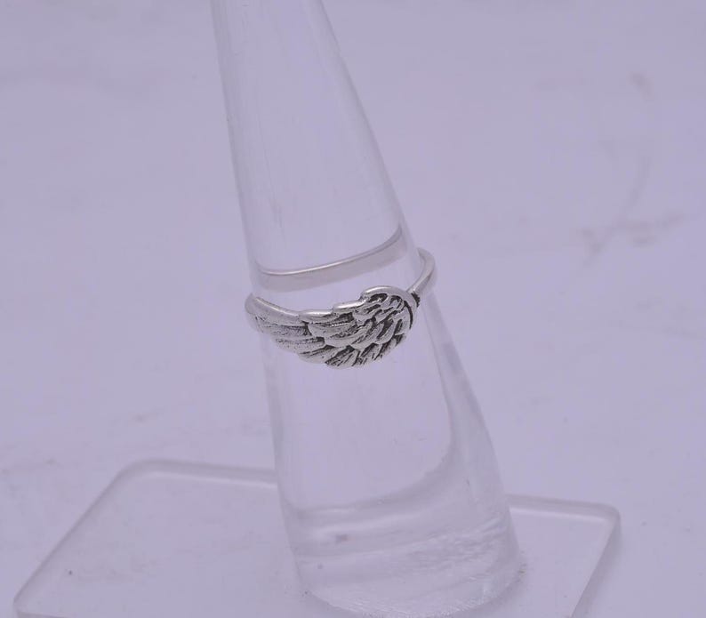 May include: A silver ring with a wing design. The ring is displayed on a clear ring stand.