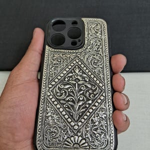 Puede incluir: Una funda de tel&eacute;fono plateada con un intrincado dise&ntilde;o floral. La funda est&aacute; hecha de metal y tiene un fondo negro.