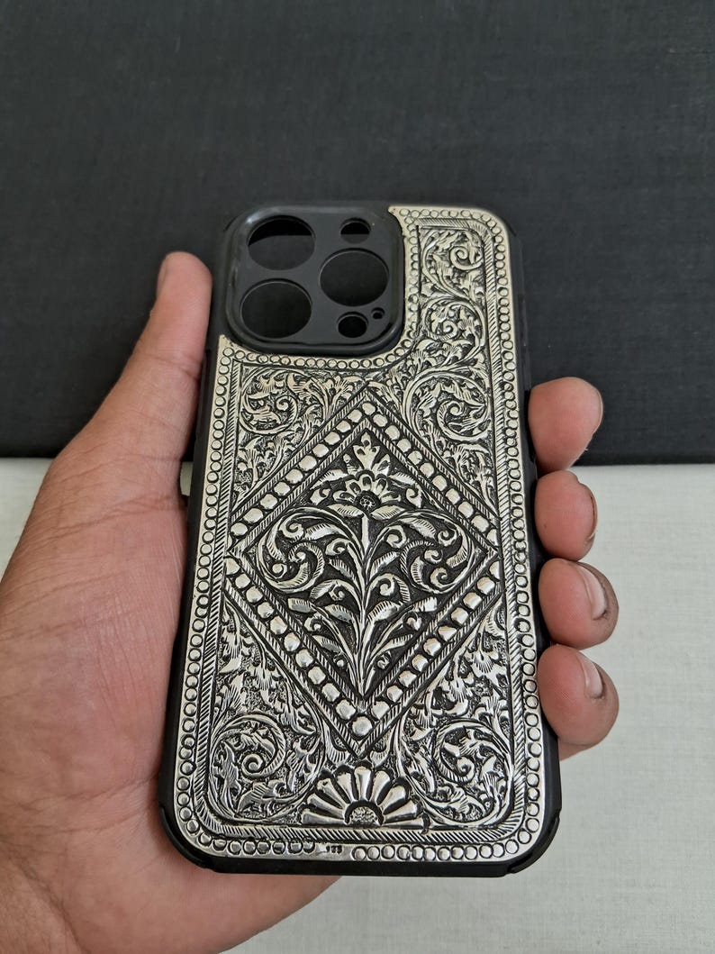 Puede incluir: Una funda de tel&eacute;fono plateada con un intrincado dise&ntilde;o floral. La funda est&aacute; hecha de metal y tiene un fondo negro.