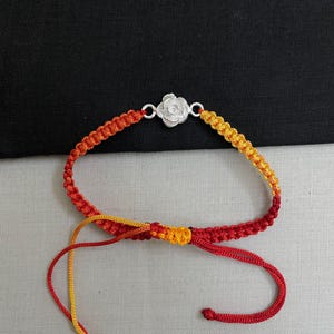 May include: A handmade bracelet featuring a woven design in shades of red, orange, and yellow. The bracelet has a silver-colored rose-shaped clasp and adjustable cords for a comfortable fit. This accessory is perfect for adding a pop of color.