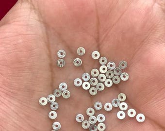 May include: A collection of small, silver-colored, flat, circular metal beads with a hole in the center. The beads are scattered across a person's open palm, with a red background visible in the upper left corner.