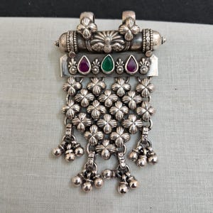 May include: A silver pendant with a detailed design featuring a floral motif and a central peacock design. The pendant has a dangling section with multiple small silver balls and a row of floral designs. The pendant is adorned with red and green gemstones.