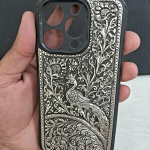 May include: A silver phone case with an intricate peacock design. The case is made of metal and has a detailed floral pattern.