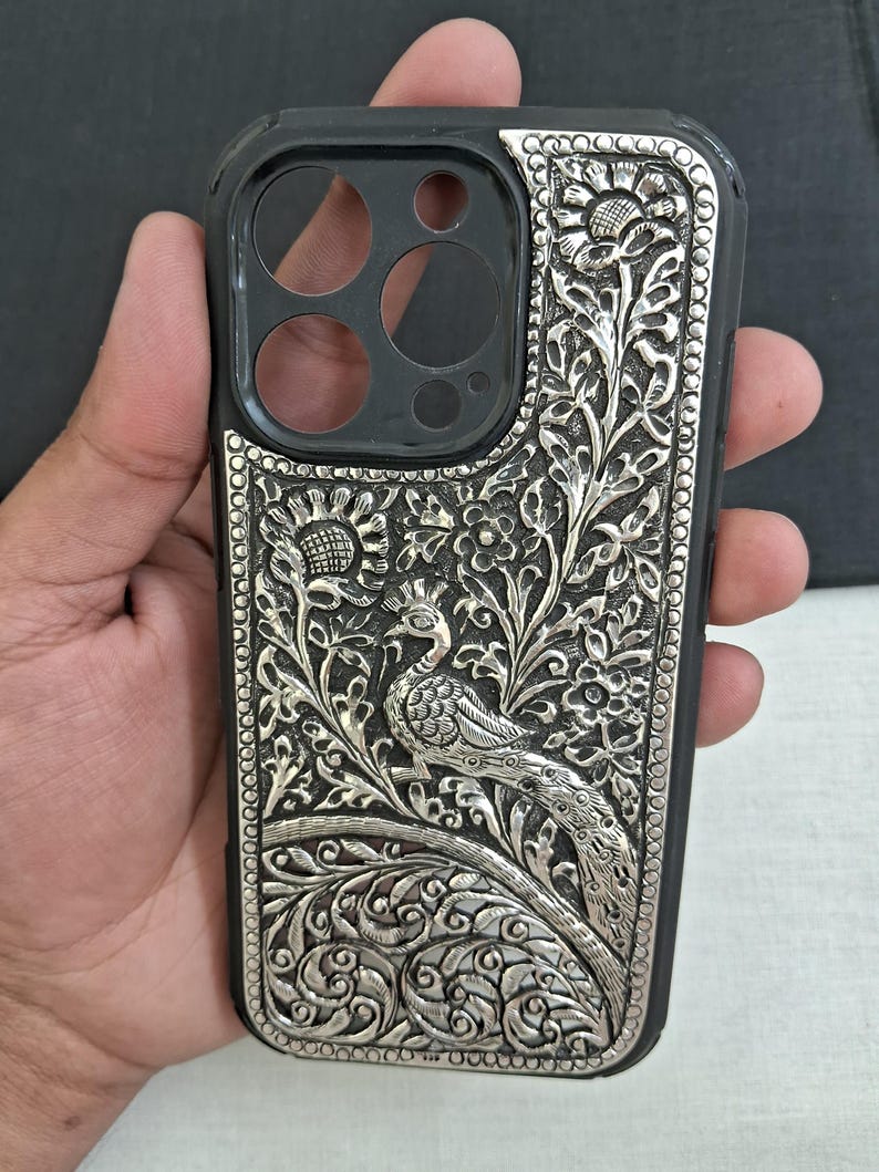 May include: A silver phone case with an intricate peacock design. The case is made of metal and has a detailed floral pattern.
