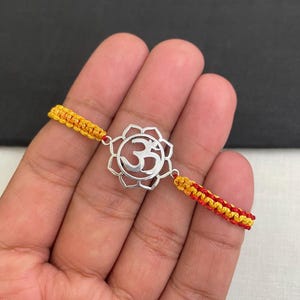 May include: Silver-toned Om symbol bracelet with a floral design. The bracelet features a woven cord in yellow and red. The Om symbol is a spiritual icon in Hinduism, Buddhism, and Jainism.