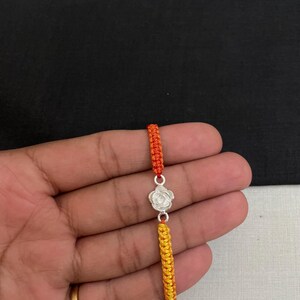 May include: A silver-colored rose charm bracelet with orange and yellow woven cords. The bracelet features a detailed rose pendant and is held against a neutral background.