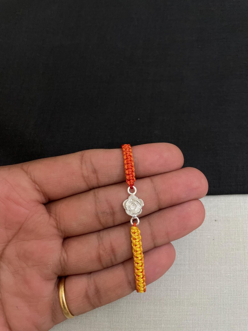 May include: A silver-colored rose charm bracelet with orange and yellow woven cords. The bracelet features a detailed rose pendant and is held against a neutral background.