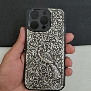 以下が含まれることがあります： 複雑な孔雀のデザインが施された銀色の携帯電話ケース。ケースは金属製で、黒色の背景。孔雀はデザインの中心にあり、花柄に囲まれている。