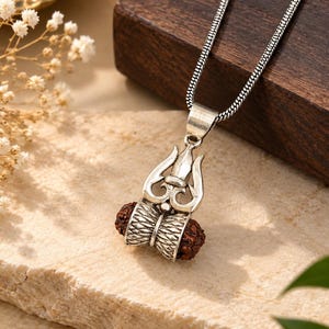 May include: A silver-toned pendant necklace featuring a trident symbol combined with a damaru drum shape. The pendant incorporates two small, textured, dark brown rudraksha beads set within the silver metal frame, suspended from a silver chain.