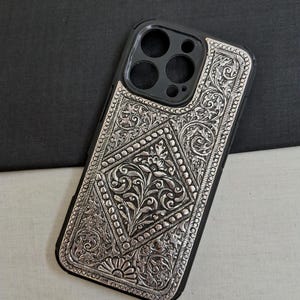 Puede incluir: Una funda de tel&eacute;fono plateada con un intrincado dise&ntilde;o floral. La funda est&aacute; hecha de metal y tiene un borde negro. El dise&ntilde;o tiene forma de diamante y presenta un patr&oacute;n floral con un dise&ntilde;o de sol en la parte inferior.