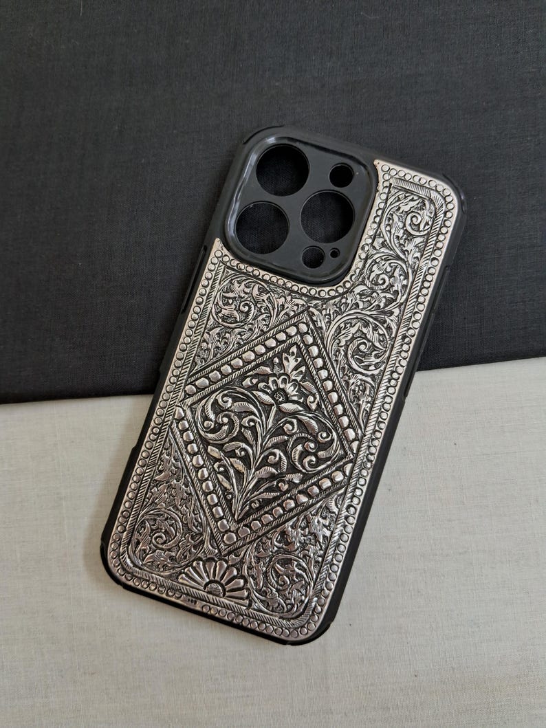 Puede incluir: Una funda de tel&eacute;fono plateada con un intrincado dise&ntilde;o floral. La funda est&aacute; hecha de metal y tiene un borde negro. El dise&ntilde;o tiene forma de diamante y presenta un patr&oacute;n floral con un dise&ntilde;o de sol en la parte inferior.