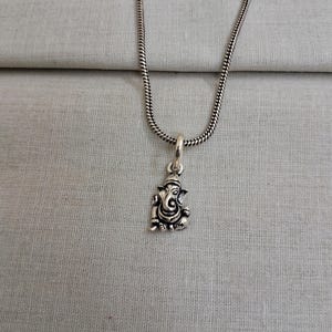 Pu&ograve; includere: Una collana in metallo color argento con un piccolo ciondolo dettagliato della divinit&agrave; Ganesha. Il ciondolo &egrave; sospeso a una sottile catena a serpente rotonda. Il metallo ha una finitura lucida con aree incavate scure.