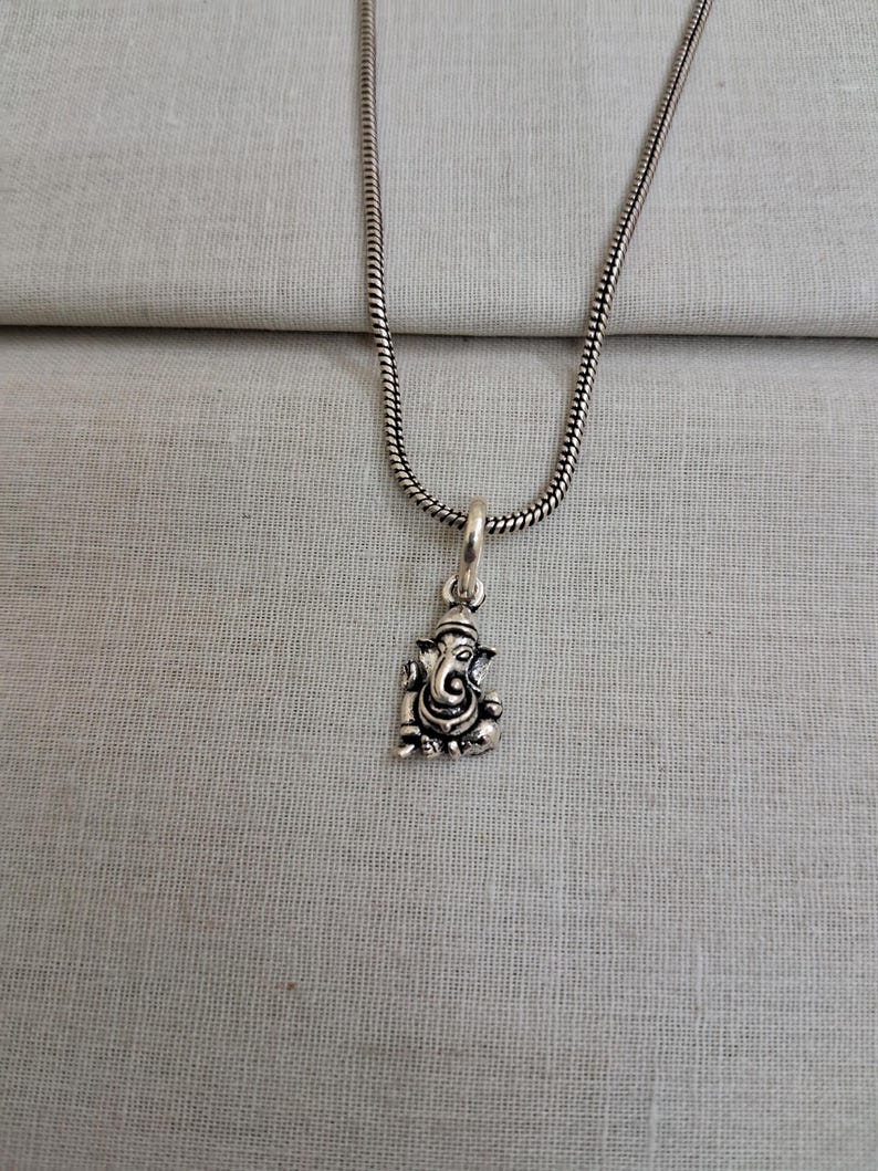 Pu&ograve; includere: Una collana in metallo color argento con un piccolo ciondolo dettagliato della divinit&agrave; Ganesha. Il ciondolo &egrave; sospeso a una sottile catena a serpente rotonda. Il metallo ha una finitura lucida con aree incavate scure.