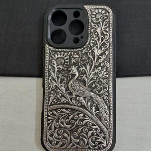 May include: A silver phone case with an intricate peacock design. The case is made of metal and has a black background.