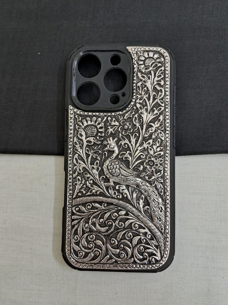 May include: A silver phone case with an intricate peacock design. The case is made of metal and has a black background.