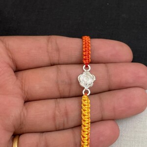 May include: A handmade bracelet featuring a silver flower charm and orange and yellow woven cord. The bracelet is held against a dark background. The cord is a vibrant mix of orange, yellow, and red.