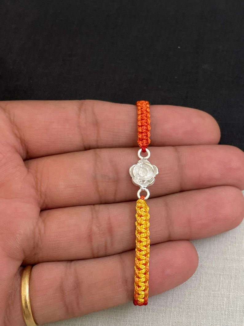 May include: A handmade bracelet featuring a silver flower charm and orange and yellow woven cord. The bracelet is held against a dark background. The cord is a vibrant mix of orange, yellow, and red.