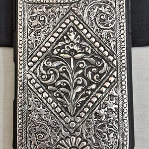 Puede incluir: Una funda de tel&eacute;fono plateada con un intrincado dise&ntilde;o floral. La funda est&aacute; hecha de metal y tiene un borde negro.