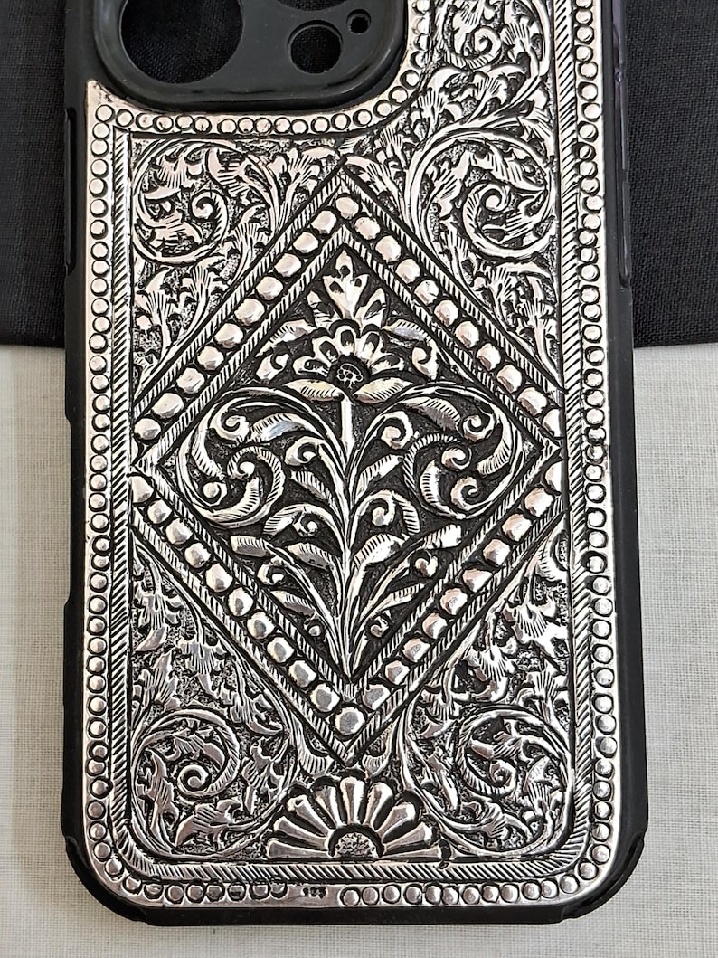 Puede incluir: Una funda de tel&eacute;fono plateada con un intrincado dise&ntilde;o floral. La funda est&aacute; hecha de metal y tiene un borde negro.