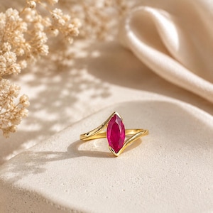 May include: A gold ring featuring a marquise-cut vibrant pinkish-red gemstone set in a bypass-style band. The metal band has a polished finish and curves around the central stone, which is positioned vertically in a secure prong setting.