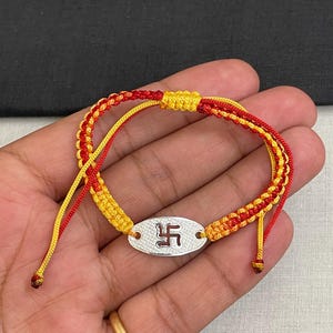 May include: A braided bracelet featuring red and yellow cord strands with an adjustable sliding knot closure. The center piece is an oval silver-toned metal charm with a textured surface and a dark red swastika symbol embossed in the middle.