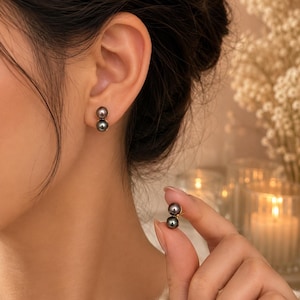 May include: A pair of earrings featuring two stacked dark grey pearls on a gold-toned post. One earring is shown worn on the earlobe, while the other is held between fingers, displaying the vertical arrangement of the two spherical pearls.