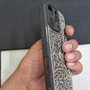 May include: A black phone case with a silver, intricate design featuring a peacock and floral patterns. The case has a raised camera bump and cutouts for the buttons and ports.