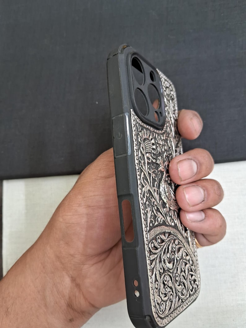 May include: A black phone case with a silver, intricate design featuring a peacock and floral patterns. The case has a raised camera bump and cutouts for the buttons and ports.