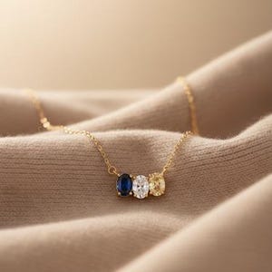 Könnte beinhalten: Eine goldene Halskette mit drei ovalen Edelsteinen: einem dunkelblauen Saphir, einem klaren Diamanten und einem gelben Saphir. Die Halskette liegt auf beigem Stoff.
