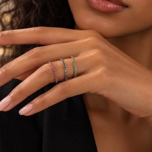 May include: Three thin metal rings worn on a finger, each featuring a continuous row of small, pavé-set gemstones. The rings are colored in vibrant pink, deep blue, and bright green, showcasing a slender band design with a polished finish.