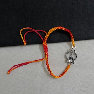 May include: A handmade bracelet featuring a braided cord in red and yellow. The bracelet has a silver-colored charm with an ornate design. The cord is adjustable with sliding knots.