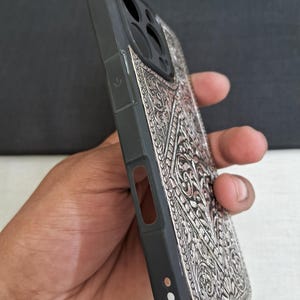 Puede incluir: Una funda de tel&eacute;fono negra con un dise&ntilde;o plateado, intrincado y en relieve. La funda tiene un recorte para el objetivo de la c&aacute;mara y un bot&oacute;n en el lateral.