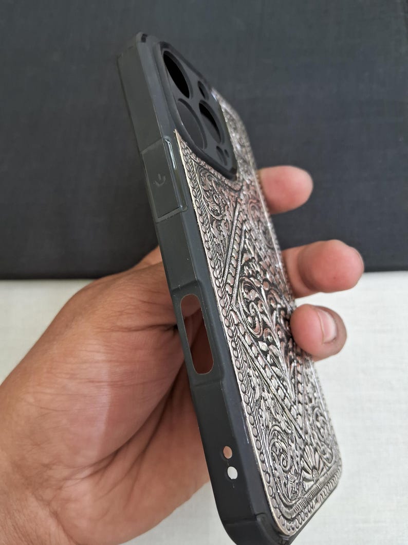 Puede incluir: Una funda de tel&eacute;fono negra con un dise&ntilde;o plateado, intrincado y en relieve. La funda tiene un recorte para el objetivo de la c&aacute;mara y un bot&oacute;n en el lateral.