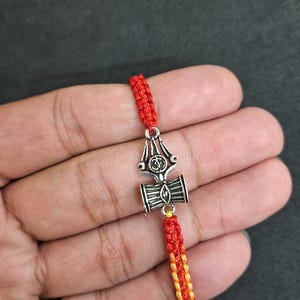Könnte beinhalten: Ein rot-gelb geflochtenes Armband mit einem silbernen Anhänger, der einen Dreizack und eine kleine Trommel zeigt. Der Anhänger ist ein Symbol von Shiva, einer hinduistischen Gottheit.