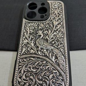 May include: A silver phone case with an intricate peacock design. The case is made of metal and has a black rubber bumper.