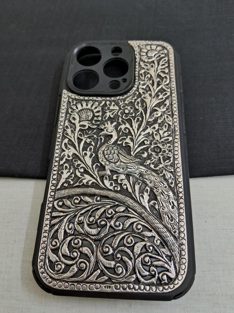 May include: A silver phone case with an intricate peacock design. The case is made of metal and has a black rubber bumper.