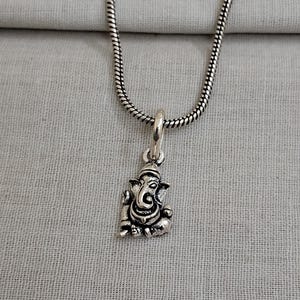 Pu&ograve; includere: Una collana in metallo color argento con un piccolo ciondolo dettagliato a forma della divinit&agrave; Ganesha. Il ciondolo &egrave; appeso a una sottile catena a serpente flessibile. Il metallo ha una finitura lucida con aree scure per i dettagli.