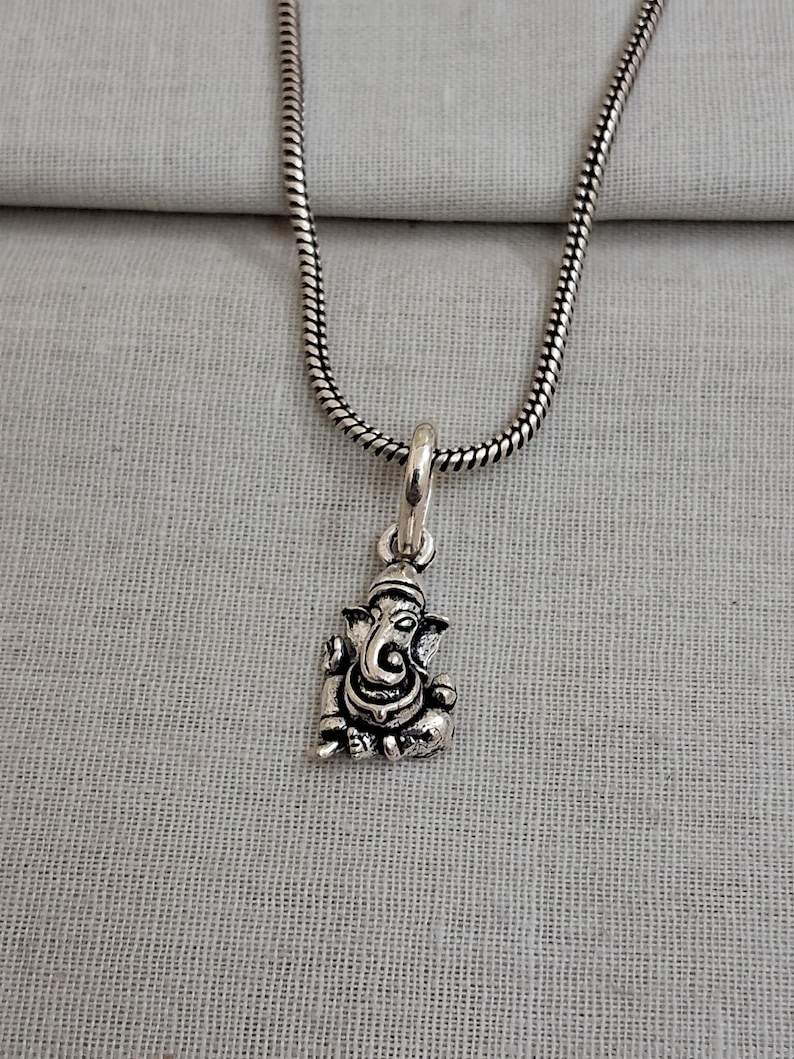 Pu&ograve; includere: Una collana in metallo color argento con un piccolo ciondolo dettagliato a forma della divinit&agrave; Ganesha. Il ciondolo &egrave; appeso a una sottile catena a serpente flessibile. Il metallo ha una finitura lucida con aree scure per i dettagli.