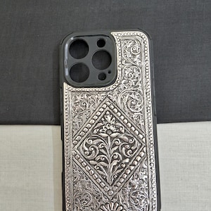 Puede incluir: Una funda de tel&eacute;fono plateada con un intrincado dise&ntilde;o floral. La funda est&aacute; hecha de metal y tiene un borde negro alrededor de los bordes.
