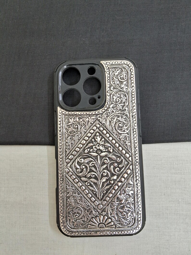 Puede incluir: Una funda de tel&eacute;fono plateada con un intrincado dise&ntilde;o floral. La funda est&aacute; hecha de metal y tiene un borde negro alrededor de los bordes.