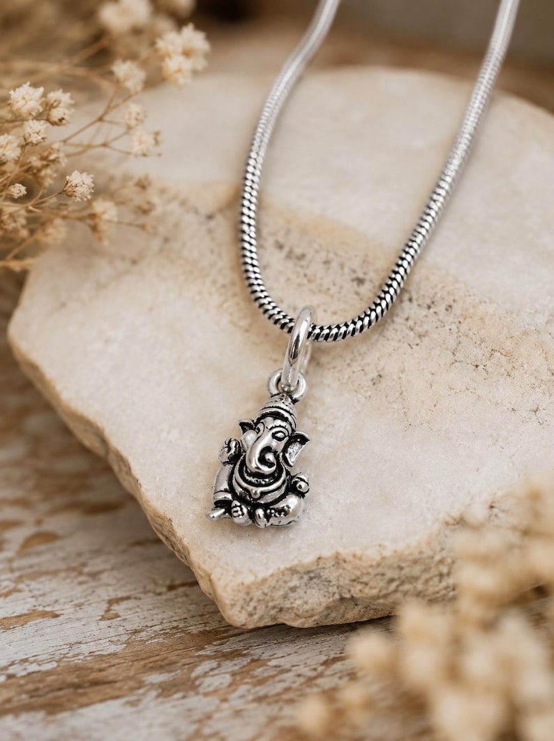Pu&ograve; includere: Un ciondolo in metallo color argento a forma della divinit&agrave; Ganesha, caratterizzato da dettagli intricati e una postura seduta. Il ciondolo &egrave; sospeso a una catena a serpente color argento abbinata, con una finitura lucida e aree incavate scure.