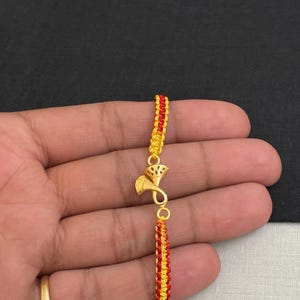 May include: A gold-colored bracelet with a trumpet-shaped charm. The bracelet features woven sections in red and yellow. The charm is a stylized trumpet with decorative elements. The bracelet is held in a hand.