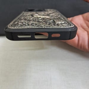 以下が含まれることがあります： 銀色の装飾的なデザインが施された黒い携帯電話ケース。ケースには、携帯電話のカメラとスピーカー用の切り抜きがあります。