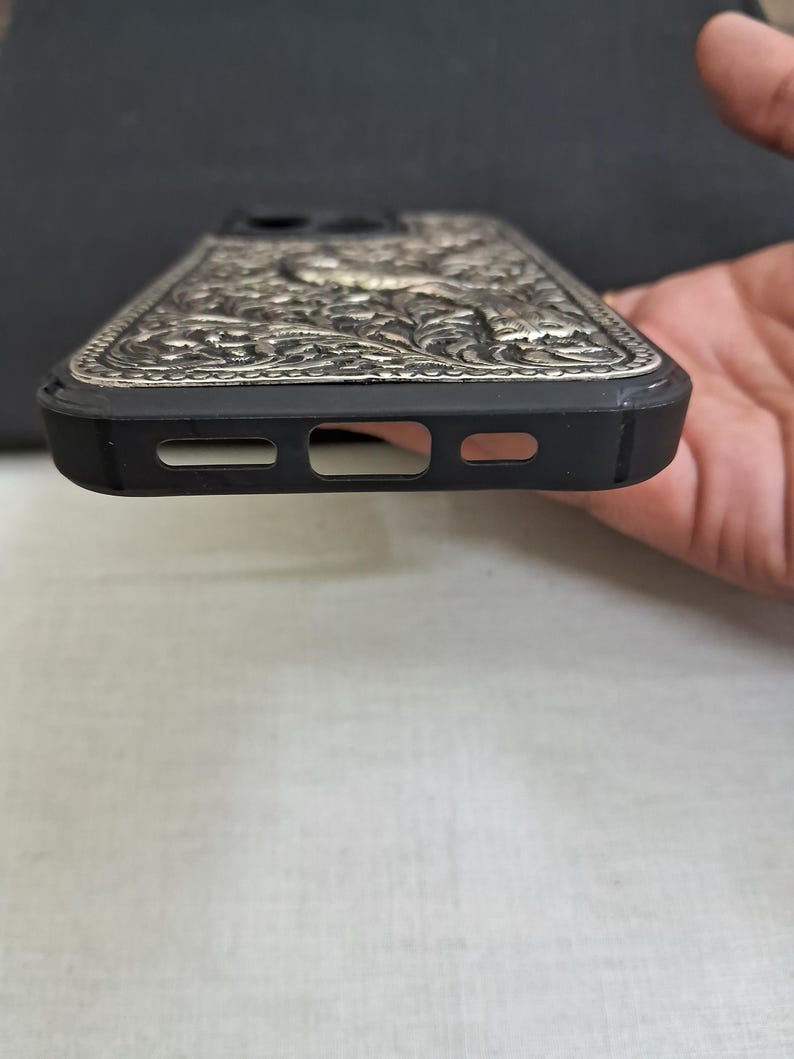 以下が含まれることがあります： 銀色の装飾的なデザインが施された黒い携帯電話ケース。ケースには、携帯電話のカメラとスピーカー用の切り抜きがあります。
