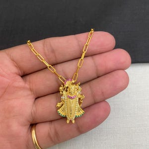 May include: A gold pendant necklace featuring a detailed figure with a crown and ornate clothing. The pendant is adorned with red, green, and white gemstones. The necklace has a delicate chain and is displayed on a hand.