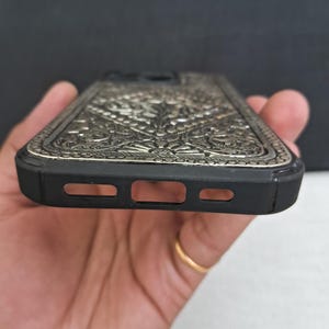 Puede incluir: Una funda de tel&eacute;fono negra con un dise&ntilde;o floral plateado intrincado. La funda tiene un borde elevado alrededor de la lente de la c&aacute;mara y un recorte para el puerto de carga.
