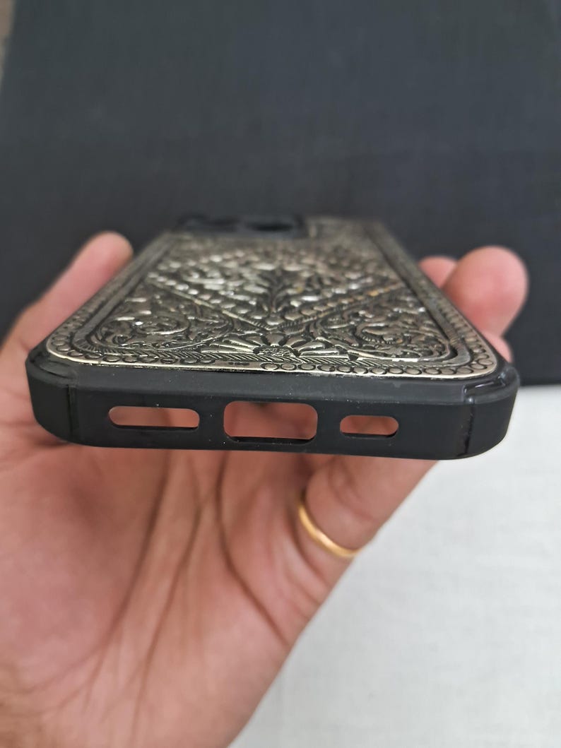 Puede incluir: Una funda de tel&eacute;fono negra con un dise&ntilde;o floral plateado intrincado. La funda tiene un borde elevado alrededor de la lente de la c&aacute;mara y un recorte para el puerto de carga.