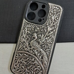 May include: A silver phone case with an intricate peacock design. The case is made of metal and has a black background. The peacock is surrounded by floral patterns.