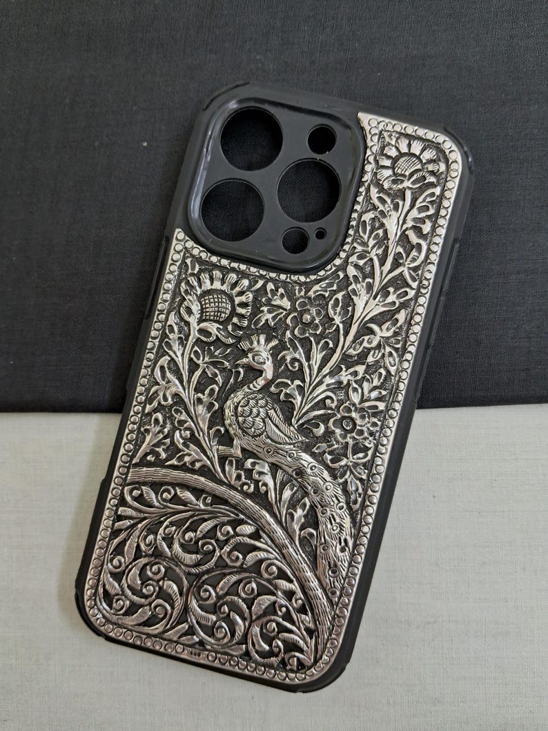 May include: A silver phone case with an intricate peacock design. The case is made of metal and has a black background. The peacock is surrounded by floral patterns.