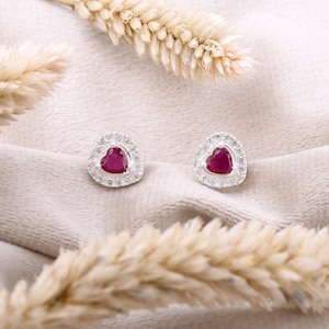 Puede incluir: Un par de pendientes de botón en forma de corazón. Cada pendiente presenta un centro de piedra preciosa roja vibrante, rodeado por un halo de pequeñas piedras transparentes. Los pendientes están colocados sobre una superficie clara y texturizada, con espigas de trigo secas en primer plano.